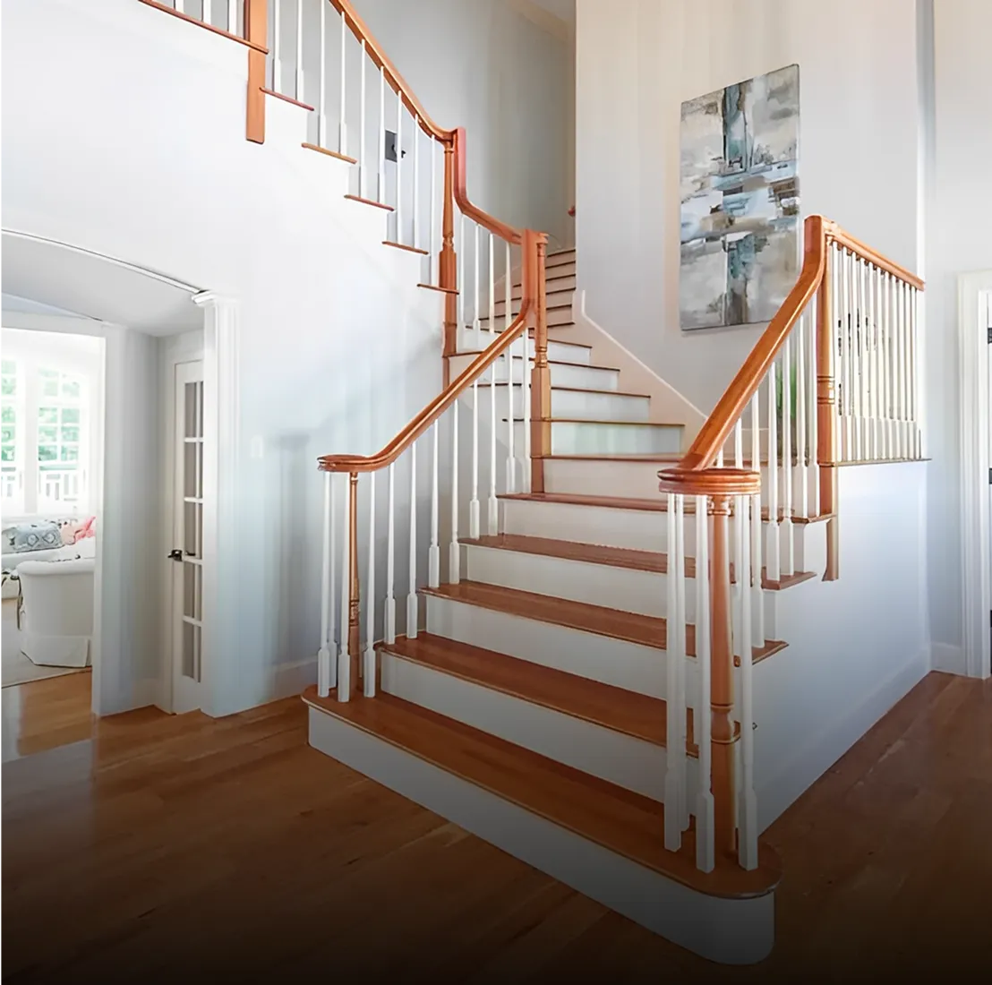 Elegant wooden staircase with white balusters and a polished handrail in a bright home interior. Artwork hangs on the wall. A doorway on the left leads to a sunlit room.