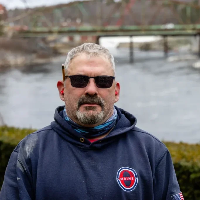 Man wearing sunglasses by a riverside bridge.